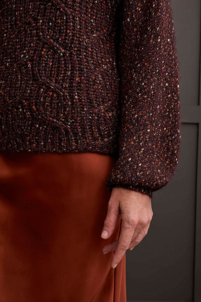 Person wearing a red patterned coat, cozy Tribal Funnel Neck Oversize Sweater in red, blue jeans, and black ankle boots stands against a dark paneled wall.