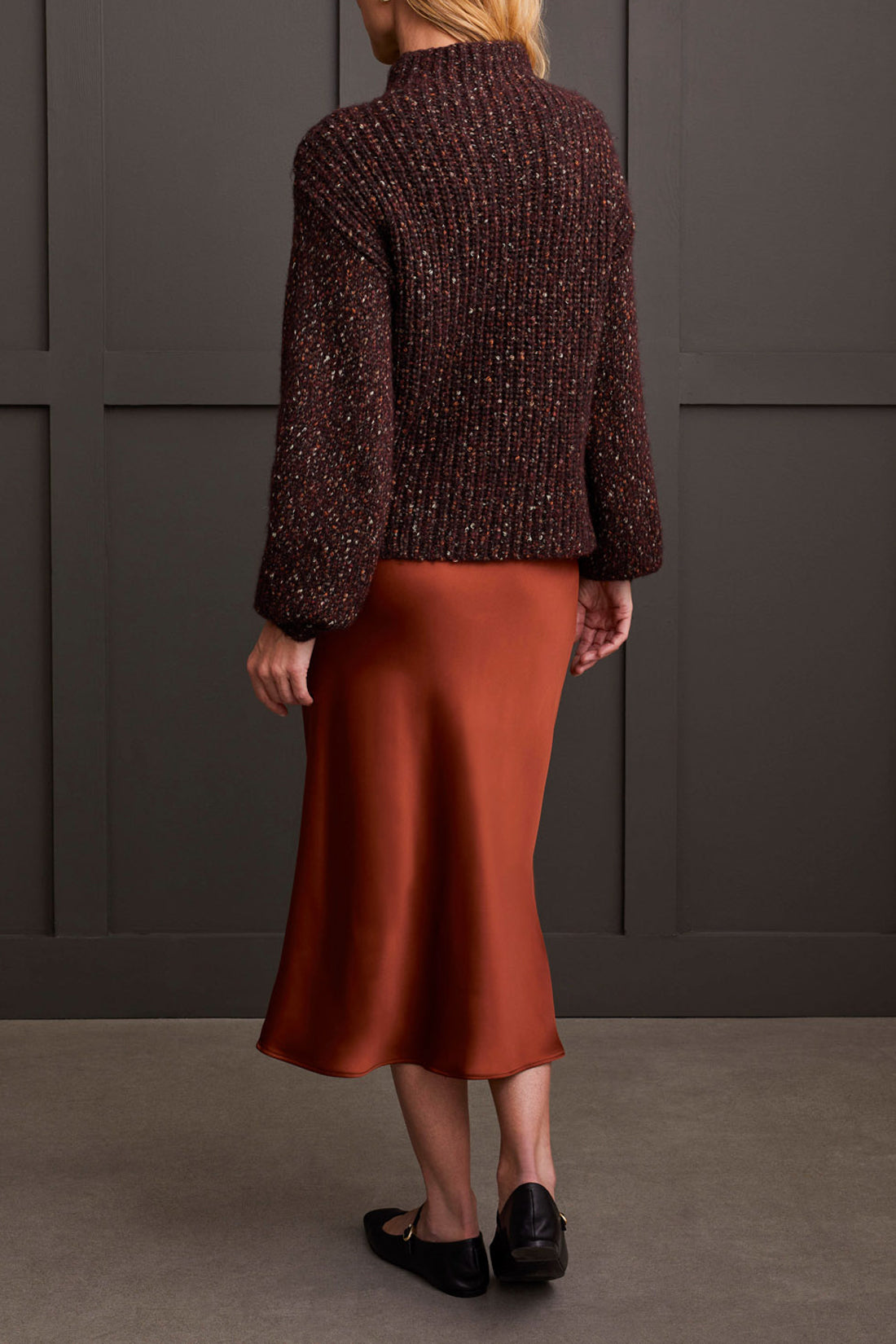 Person wearing a red patterned coat, cozy Tribal Funnel Neck Oversize Sweater in red, blue jeans, and black ankle boots stands against a dark paneled wall.