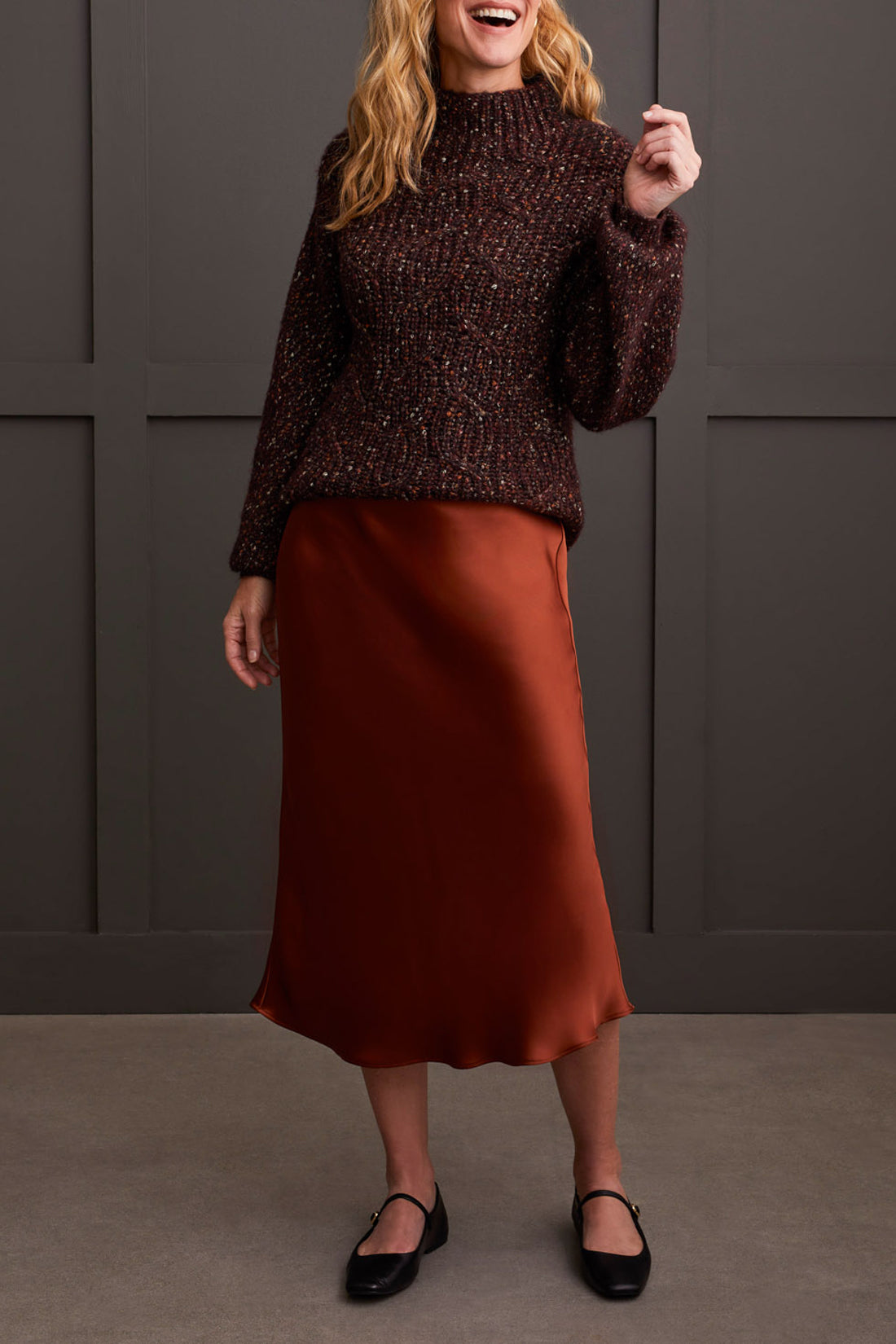 Person wearing a red patterned coat, cozy Tribal Funnel Neck Oversize Sweater in red, blue jeans, and black ankle boots stands against a dark paneled wall.