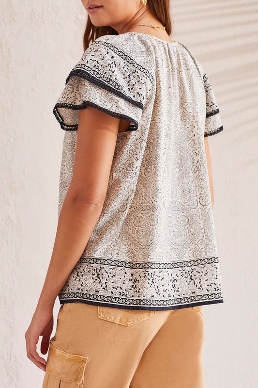 A woman facing away, wearing a Tribal Flutter Short Sleeve Blouse and beige trousers, standing against a textured wall.