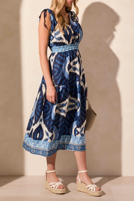 A woman wears the Tribal MAXI DRESS WITH SHOULDER TIES in blue and white, facing away while carrying a beige handbag and sporting white wedge sandals.