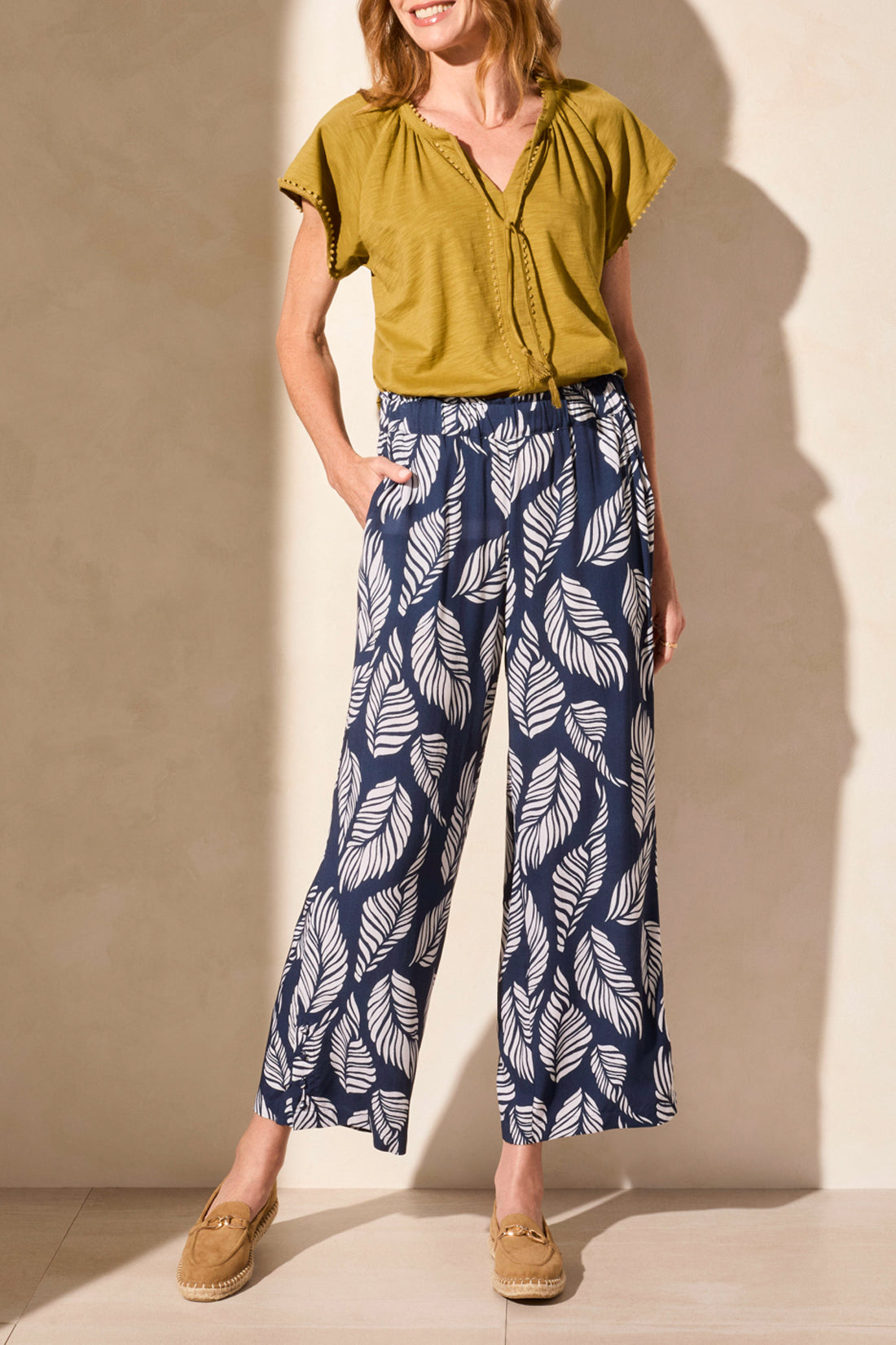 Person wearing Tribal’s PULL ON ANKLE PANT WITH HEM VENT & BUTTONS in navy with a white leaf print, styled with a green top and tan loafers, standing against a neutral background.