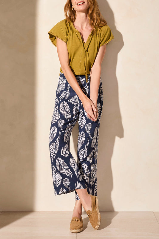 Person wearing Tribal’s PULL ON ANKLE PANT WITH HEM VENT & BUTTONS in navy with a white leaf print, styled with a green top and tan loafers, standing against a neutral background.
