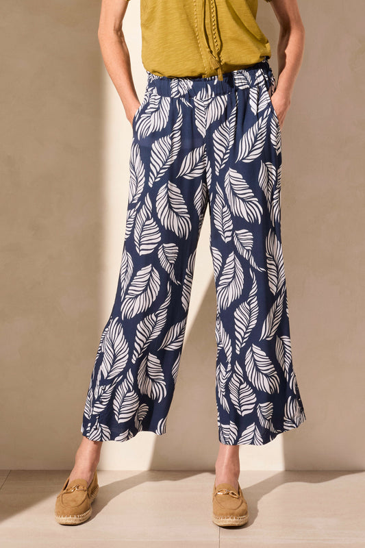 Person wearing Tribal’s PULL ON ANKLE PANT WITH HEM VENT & BUTTONS in navy with a white leaf print, styled with a green top and tan loafers, standing against a neutral background.