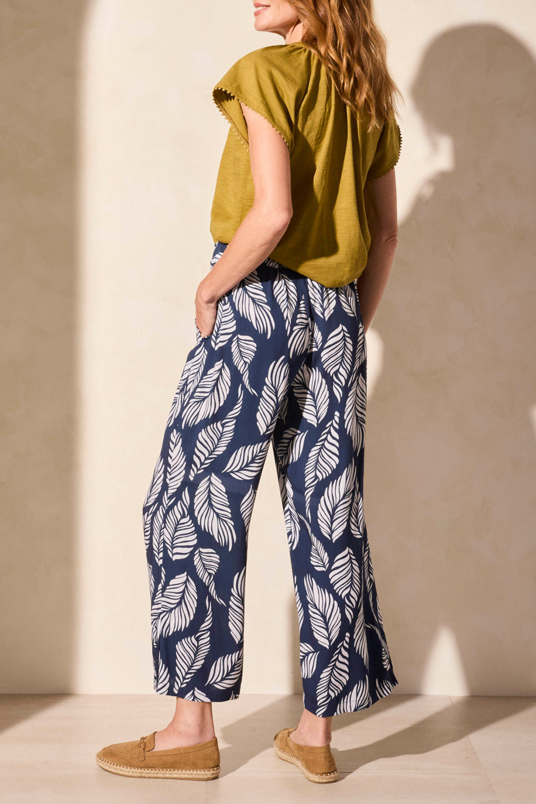 Person wearing Tribal’s PULL ON ANKLE PANT WITH HEM VENT & BUTTONS in navy with a white leaf print, styled with a green top and tan loafers, standing against a neutral background.