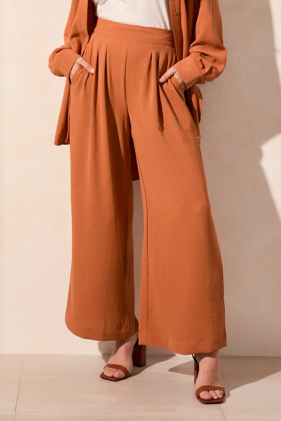 A woman stands against a neutral background wearing a rust-colored button-up shirt, a white top, brown heeled sandals, and Tribal’s FLOWY PANT WITH BLACK ELASTIC WAISTBAND.