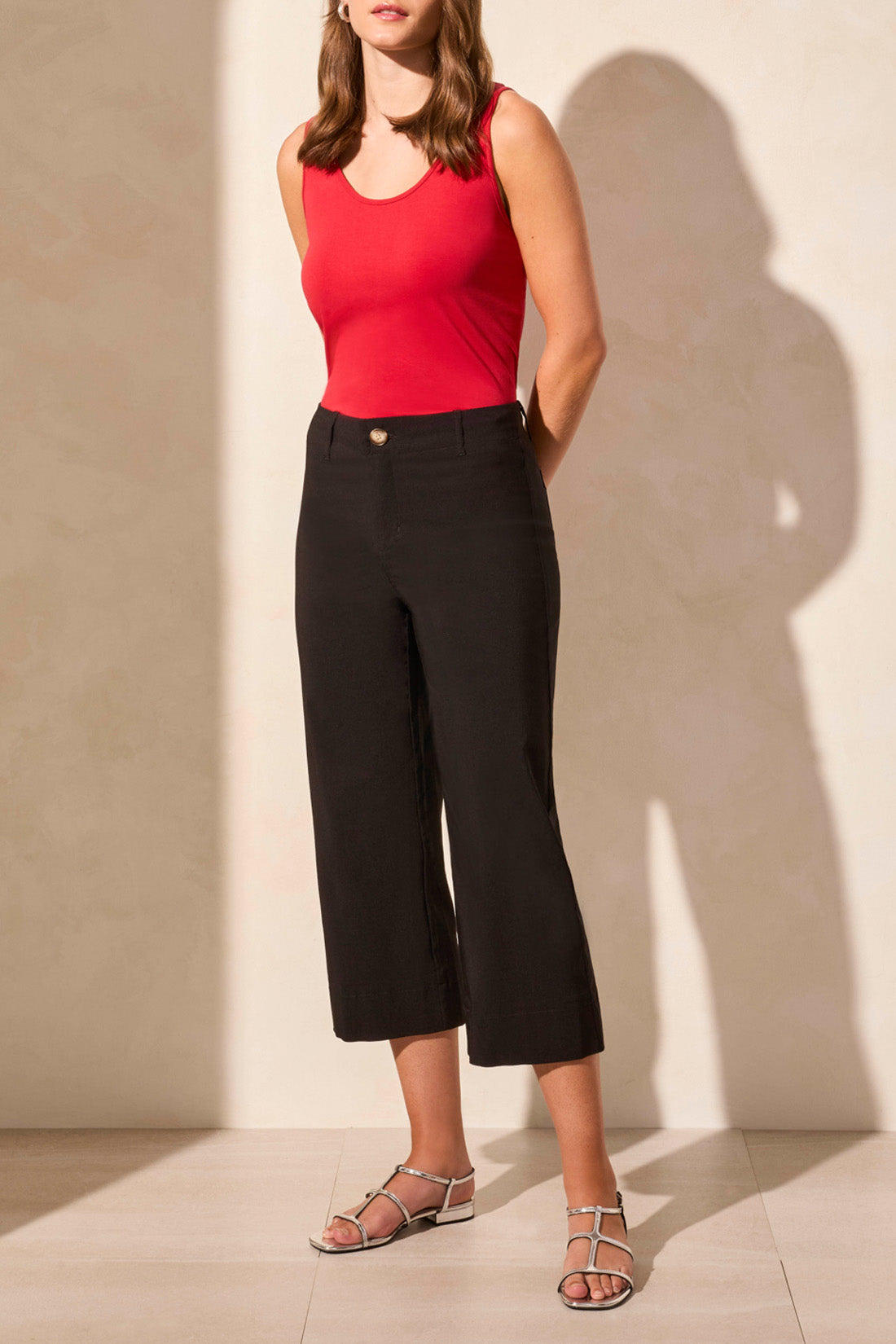 A person in a red sleeveless top, FLOWY HIGH RISE TWILL CAPRI PANT by Tribal, and sandals stands in a well-lit room with light-colored walls and floor, facing away.