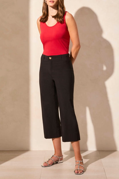 A person in a red sleeveless top, FLOWY HIGH RISE TWILL CAPRI PANT by Tribal, and sandals stands in a well-lit room with light-colored walls and floor, facing away.