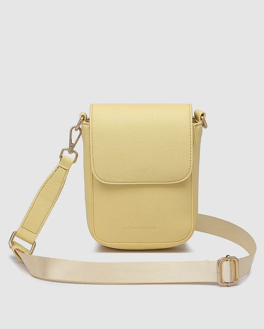 A woman indoors wears a sleeveless pink and orange dress, carrying the Louenhide Cuba Phone Bag—a pale yellow vegan leather crossbody with a wide strap—over her shoulder.