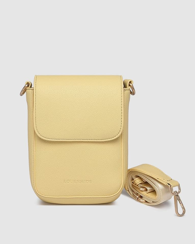 A woman indoors wears a sleeveless pink and orange dress, carrying the Louenhide Cuba Phone Bag—a pale yellow vegan leather crossbody with a wide strap—over her shoulder.