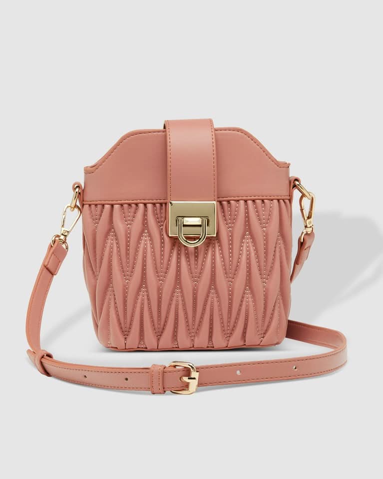 A woman outdoors holds the Louenhide Layla Crossbody Bag, a pink vegan leather mini bucket bag, paired with a one-shoulder striped top and white high-waisted pants.