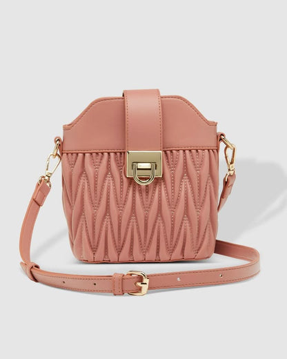 A woman outdoors holds the Louenhide Layla Crossbody Bag, a pink vegan leather mini bucket bag, paired with a one-shoulder striped top and white high-waisted pants.