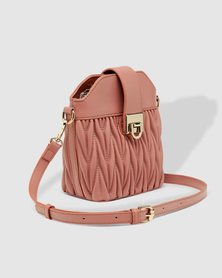 A woman outdoors holds the Louenhide Layla Crossbody Bag, a pink vegan leather mini bucket bag, paired with a one-shoulder striped top and white high-waisted pants.